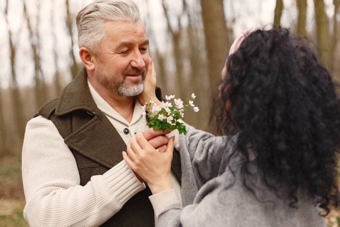 An older couple among bare trees, one offering the other a sprig of white blossom