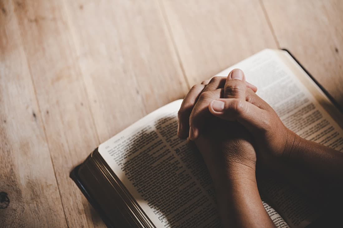 Hands held open in prayer with light falling across a worn Bible
