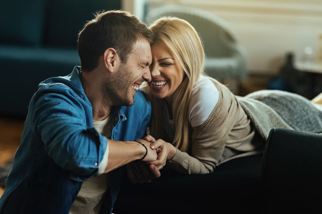A husband and wife laughing together on the living room floor, holding hands