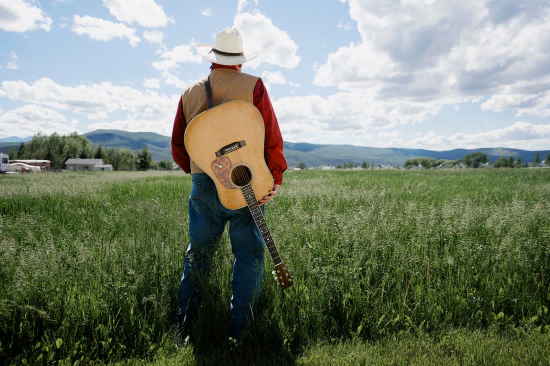 A man in a cowboy hat standing in a field with an acoustic guitar on his back