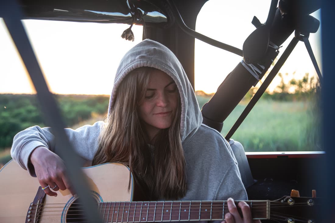 A woman playing an acoustic guitar in the back of a van at sunset